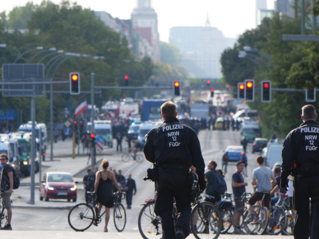 Zwei Polizisten stehen an der Seite einer Straße neben einer Gruppe von Radfahrern, mit Fahrzeugen, Verkehrszeichen, Bäumen, Gebäuden und einem klaren blauen Himmel im Hintergrund.