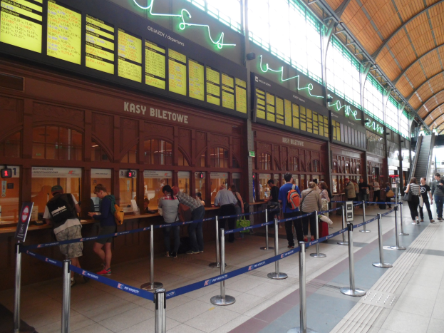 Menschen stehen in einer Schlange an einem Bahnhof mit Taschen, Stangen mit Bändern, Informationsschildern, Deckenbeleuchtung und Treppen mit Geländern in der Nähe einer Ticketstation.