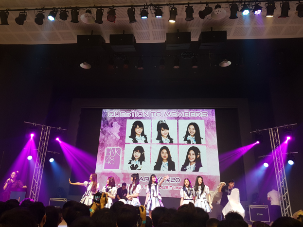 Eine Gruppe von Menschen, die auf einer hell erleuchteten Bühne performen, mit einer Menge darunter, die einen Sänger mit Mikrofon und einen Musiker mit Instrument zeigt, sowie einen großen Bildschirm mit Bildern der K-Pop-Gruppe BK48 im Hintergrund.