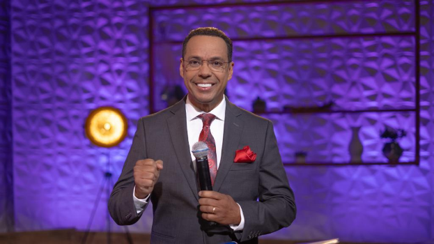 Stephen Colbert in Anzug, Krawatte und Brille auf einer Bühne mit Mikrofon stehend, mit einer Wand, Ständer, Licht und verschiedenen Gegenständen dahinter.