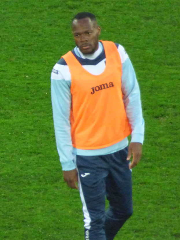 Ein Football-Spieler in einem orangen T-Shirt und blauen Hosen, der entschlossen über ein grünes Feld läuft.