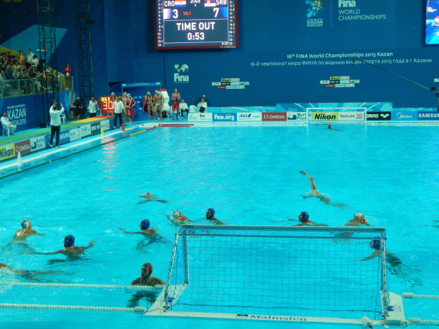Gruppe von Athleten beim Wasserballspielen in einem Pool während der 2015 FINA World Championships in Kazan, mit Zuschauern und Eventbeschilderung im Hintergrund.