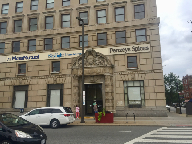 Großes Gebäude mit "Massachusetts Mutual Financial Group"-Schriftzug, parkende Autos davor, Straßenlaternen, fahrende Autos, Fußgänger auf dem Gehweg, Bäume, Topfpflanzen und bewölkter Himmel.