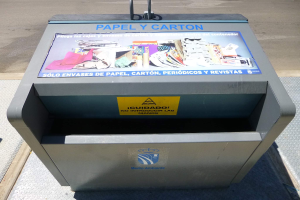 Ein Metallmüllcontainer am Straßenrand mit einem "Papel y Carton"-Plakat und fahrende Fahrzeuge im Hintergrund.