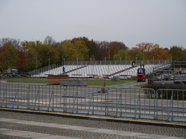 Großes Stadion mit Metallgeländern, umgeben von Bäumen unter einem klaren blauen Himmel, mit Menschen und Fahrzeugen im Vordergrund auf grünem Rasen.