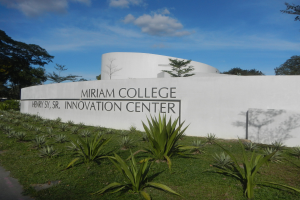 Außenansicht des Miriam College Innovation Center mit einer textbedeckten Wand, umgeben von Grün, einem Weg, Bäumen und einem bewölkten Himmel.