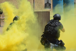 Zwei Polizisten in Einsatzausrüstung mit gelbem Rauch aus dem Mund, mit Helmen und Schutzausrüstung vor einer Wand mit einer Tafel und anderen Gegenständen, mit dem Boden unten sichtbar.