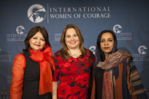 Drei Frauen stehen zusammen vor einem Banner mit der Aufschrift "Internationale Frauen des Mutes", sie lächeln und scheinen feierlich.