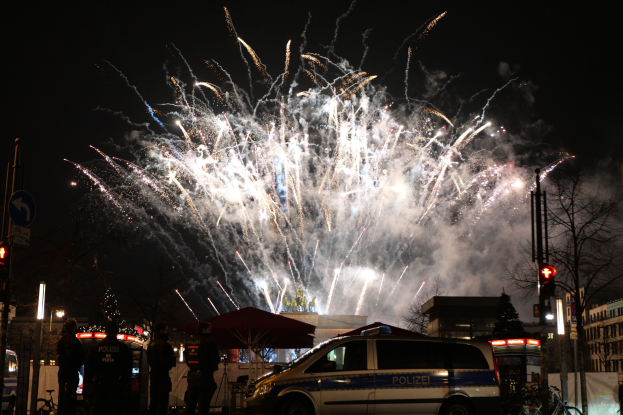 Polizeiauto vor einer Menschenmenge während einer Silvesterfeier in Berlin geparkt, mit farbigen Feuerwerkskörpern, die den Himmel erleuchten und die Stadtlichter im Hintergrund.