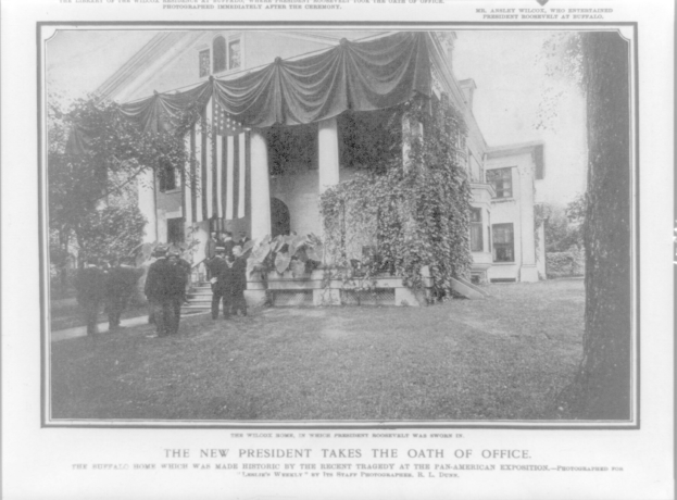 Schwarzes und weißes Foto einer Gruppe von Menschen, die vor einem Gebäude mit Fenstern, Säulen, einem Vorhang, Pflanzen und Bäumen stehen, mit Text unten: "Der neue Präsident leistete den Amtseid."