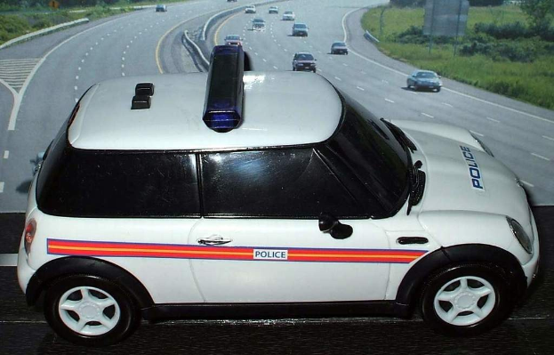 Weißer Polizeiwagen mit "Polizei" Aufschrift auf einer Straße außerhalb der Stadt, umgeben von fahrendem Verkehr, Gras und Bäumen auf beiden Seiten.