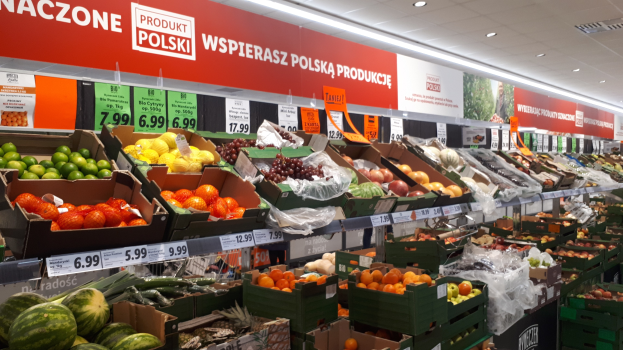 Ein Geschäft mit verschiedenen Obst- und Gemüsesorten in Kartons mit Preisschildern, Wandtafeln mit Text und Bildern und Deckenbeleuchtung.