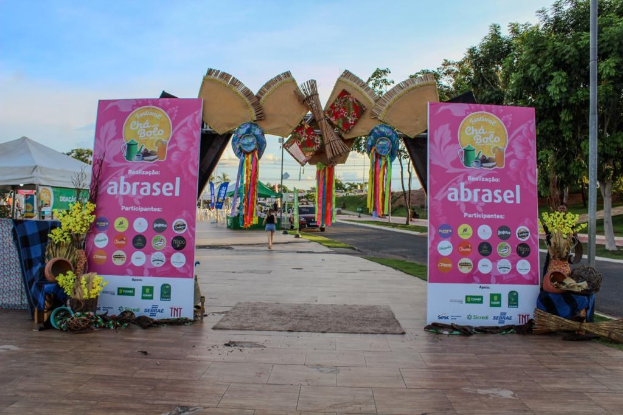Gruppe von Menschen vor einem "Abrasel"-Schild auf einem Festival mit Bannern, einem sitzenden Menschen, einem Besen, Topfpflanzen, einem Tisch mit Gegenständen, einem Zelt, Polen, Gras, Baumrinde, dekorativen Elementen, einer Straßenlaterne, Fahrzeugen und Wolken am Himmel.