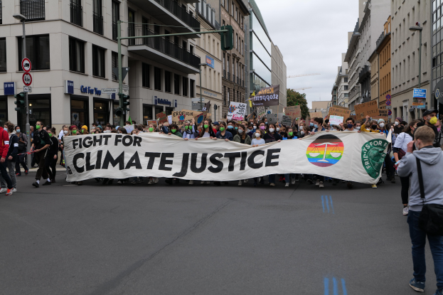 Eine Gruppe von Menschen marschiert mit einem Banner, auf dem "Kampf für Klimagerechtigkeit" steht, eine Straße entlang, mit Gebäuden, Schildern, Verkehrsampeln und Bäumen an der Straße und einem bewölkten Himmel.