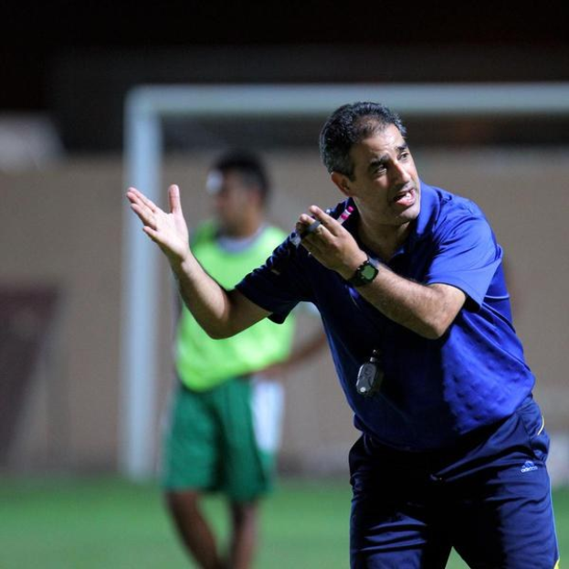 Ein Mann in einem blauen T-Shirt und Hose steht auf einem Fußballfeld und hält ein Mikrofon in der Hand, während er in Richtung der Spieler gestikuliert; im Hintergrund sind eine Person in einem grünen Kleid, ein Torpfosten und eine Wand zu sehen.