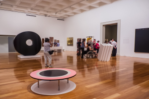 Eine Gruppe von Menschen in einer Kunstgalerie mit einer Person im Rollstuhl in der Mitte, umgeben von Rahmenfotos an den Wänden und Deckenleuchten, mit einem schwarzen Installationsobjekt links.