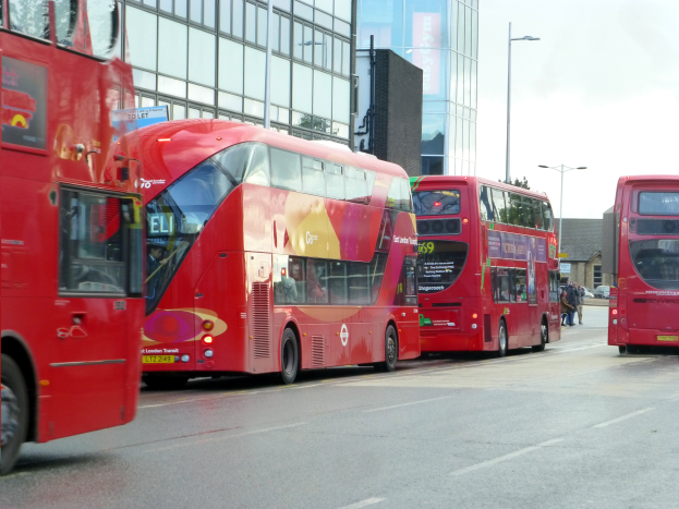 Drei rote Doppeldeckerbusse fahren eine Straße mit hohen Gebäuden entlang, mit Menschen auf dem Gehweg und Laternenmästen.