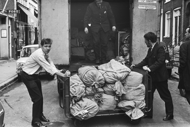 Eine Gruppe von Männern entlädt Taschen von einem Lastwagen, der auf der Seite einer Straße geparkt ist, mit einem vorbeifahrenden Auto und Gebäuden mit Fenstern, Treppen mit Geländern und Texttafeln im Hintergrund, alles in Schwarz-Weiß dargestellt.