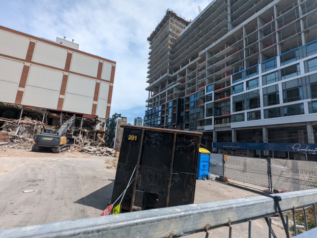 Großes Gebäude mit Schutt davor, umgeben von einem Metallzaun, einem Container, Pflanzen, Bäumen, Pfählen, einem Schild, Fahrzeugen, Steinen und einem bewölkten Himmel, was auf eine Baustelle für ein neues Wohngebäude hinweist.