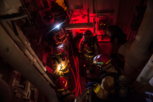 Feuerwehrleute in Helmen und Sauerstoffflaschen stehen in einem schwach beleuchteten Raum mit Rohren auf beiden Seiten, mit einer Maschine und anderen Gegenständen im Hintergrund.