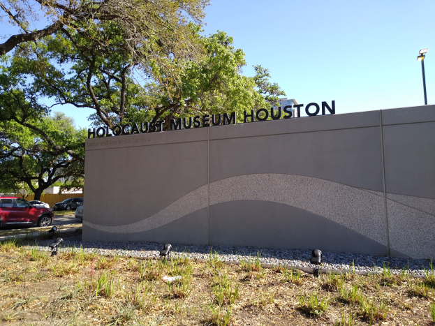 Außenansicht des Houston Holocaust Museums mit eingravierter Textwand, Gedenksteine, Grünanlage, Straße mit Fahrzeugen, Straßenlaterne und bewölktem Himmel.
