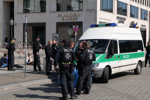 Eine Gruppe von Polizisten steht vor einem Starbucks-Café, rechts daneben parkt ein Van, links stehen und sitzen Menschen, im Hintergrund ein Gebäude mit Fenstern, ein Schild, ein Laternenpfahl und eine Absperrung.