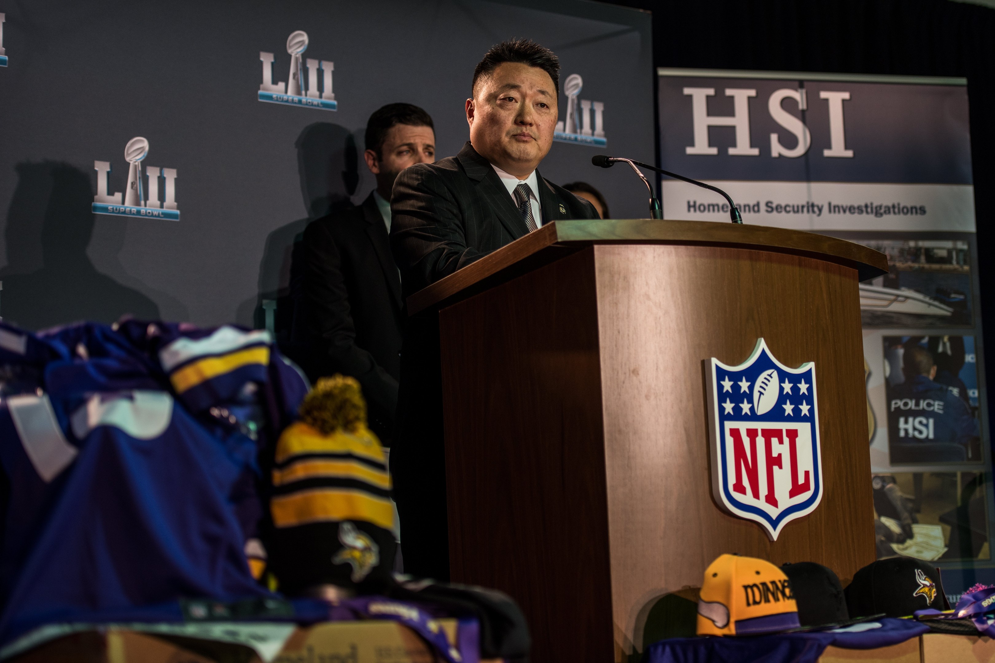 Ein Mann in einem schwarzen Blazer, einem wei├čem Hemd und einer schwarzen Krawatte spricht an einem Podium mit einem Mikrofon und Logo, gewandt einer Menge, mit blauer und gelber Merchandise zu seiner Linken und einem "Super Bowl LII"-Banner im Hintergrund.