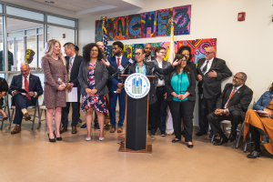Gruppe von Menschen vor einem Podium mit einem Mikrofon und einer Tafel mit der Aufschrift "Sancho Stadtverordnetenvorsteher tritt dem Stadtrat bei", mit sitzenden und stehenden Teilnehmern, Flaggen, einem Feuerlöscher und Deckenleuchten im Hintergrund.