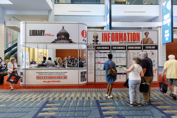 Eine Gruppe von Menschen auf dem Nationalen Buchfestival, einige sitzend und einige stehend, vor einer Bude mit Bannern, einem Anzeigebildschirm, einer Treppe, einem Geländer und einem Dach mit Deckenbeleuchtung.