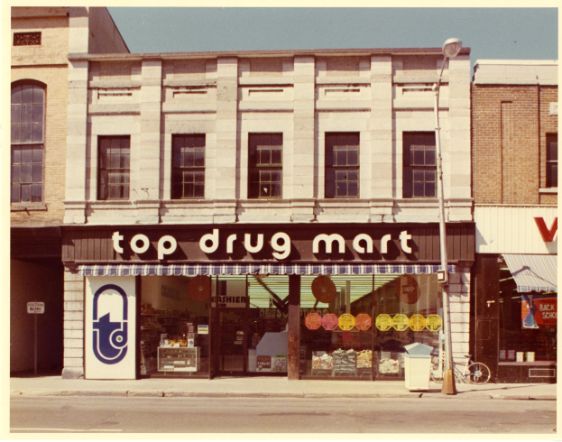 Straßensicht eines Top-Drogenmart-Geschäfts mit einem Gebäude, das Fenster und ein Schild hat, einem Straßenschild, einem geparkten Fahrrad, einem Müllcontainer und einem bewölkten Himmel.