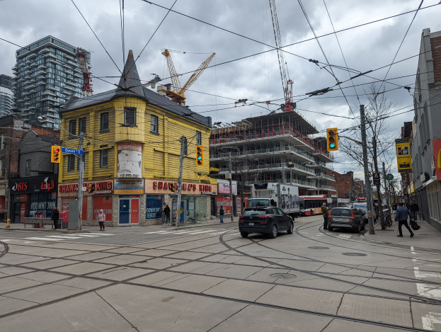 Eine Straßenszene in der Stadt mit einem gelben Gebäude im Bau, Autos, Fußgängern, Strommasten, Ampeln, Gebäuden, Schildern, Bäumen, Kränen und einem bewölkten Himmel.