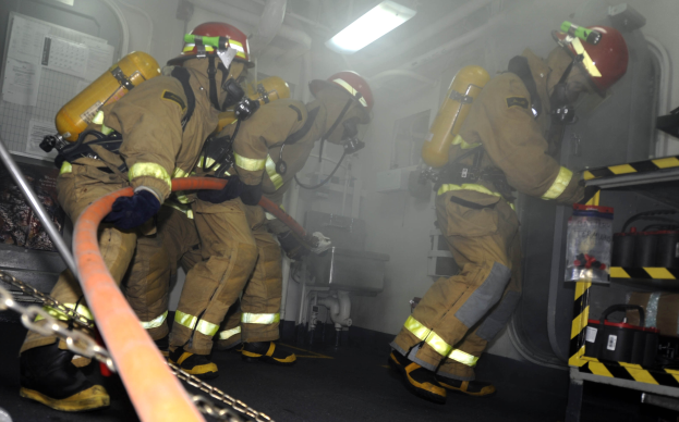 Feuerwehrleute in Schutzausrüstung und Helmen arbeiten neben einem Feuerwehrauto, mit Equipment-Regalen und Hintergrund-Infrastruktur.