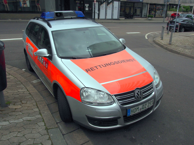 Polizeiauto am Straßenrand geparkt mit einem Müllcontainer auf dem Gehweg links, Gebäude und Bäume im Hintergrund.