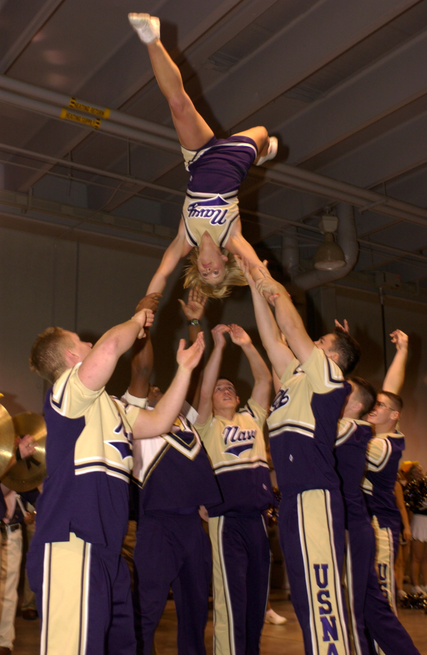 Eine Gruppe von Cheerleadern führt einen Stunt in einer Turnhalle durch, wobei eine Frau in einem Handstand in der Mitte steht, umgeben von anderen, einige halten Instrumente, und eine Wand mit Rohren und Deckenleuchten im Hintergrund.