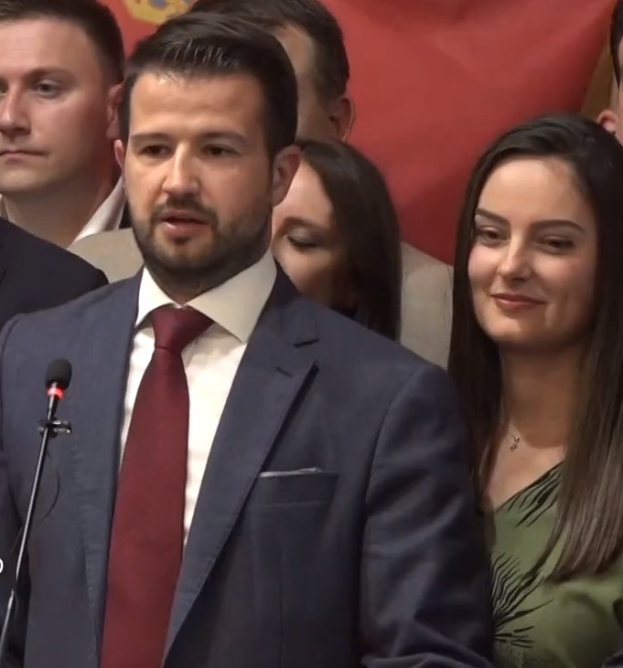 Ein Mann im Anzug spricht an einem Podium mit einem Mikrofon und blickt auf eine Menge hinter einem Banner, möglicherweise spricht er über ein Regierungsverbot von Social Media.