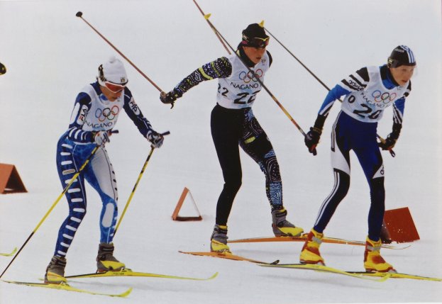 Drei Skifahrer in buntfarbigen Anzügen und Helmen rasen eine schneebedeckte Piste hinunter, jeder hält Ski-Stöcke in den Händen.