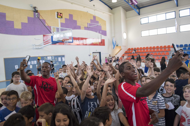Gruppe von Kindern vor einem Basketballfeld, einige halten Mobiltelefone, mit einer Tafel, Uhr, Torpfosten, Basketballnetz, Deckenleuchten, Stühlen und Fenstern im Hintergrund.