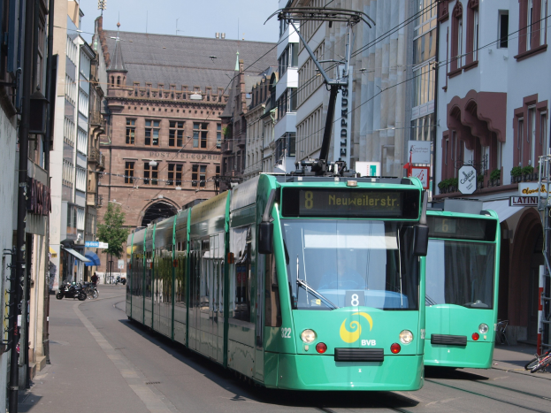 Zwei grüne Straßenbahnen fahren eine Stadtstraße mit hohen Gebäuden, Fahrrädern auf dem Gehweg, Fußgängern auf der Straße, einem Baum im Hintergrund und einem klaren blauen Himmel.