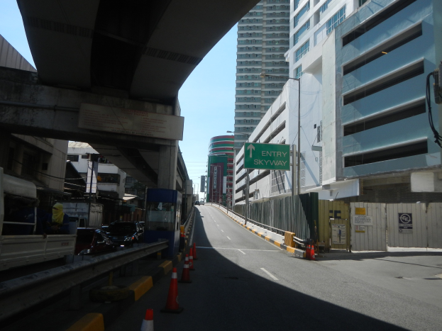 Stadtstraße mit Verkehrskegeln, Fahrzeugen, einer Brücke auf der linken Seite, Gebäuden mit Namensschildern, Laternenpfählen und Himmel im Hintergrund.