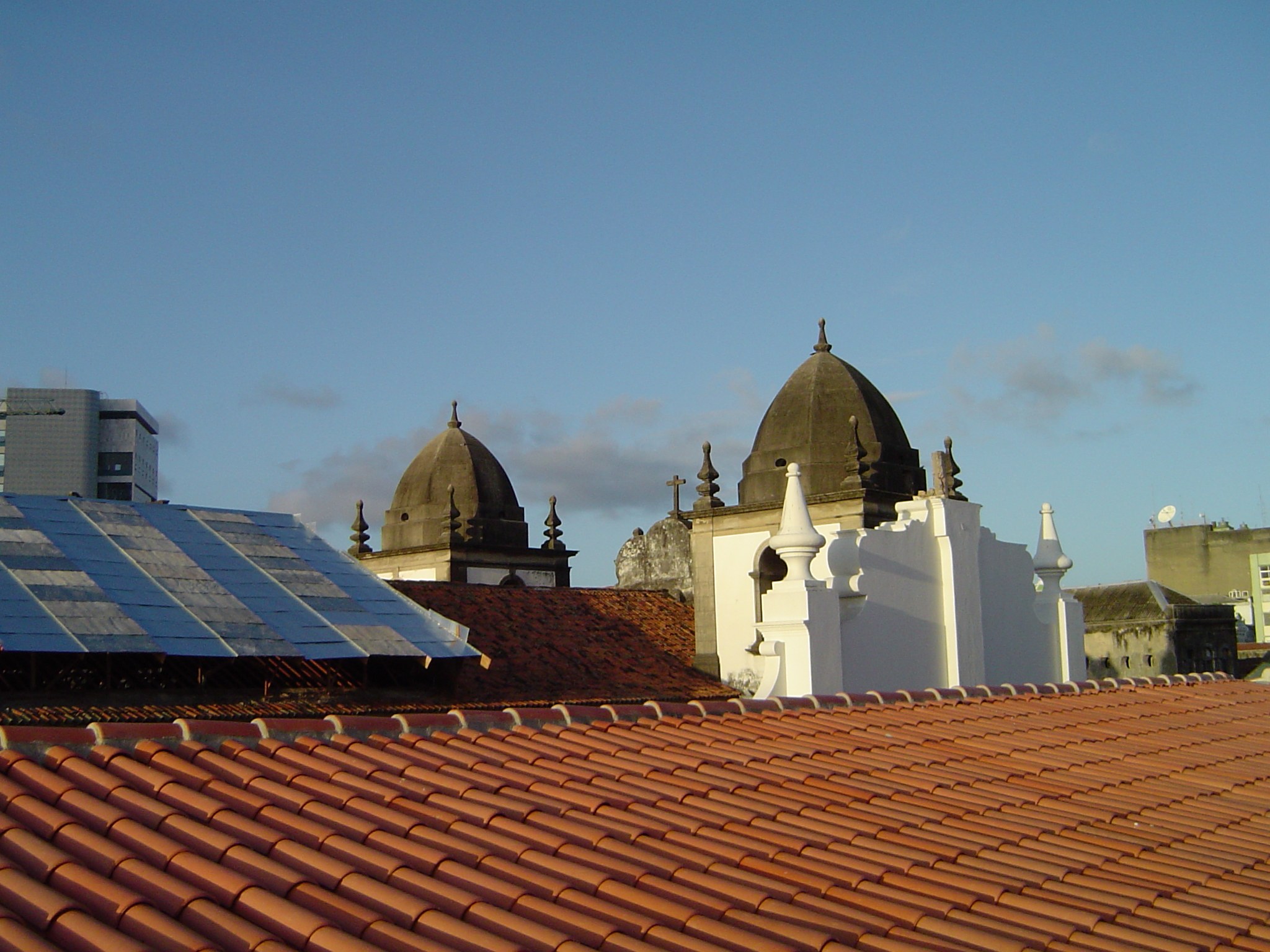 Stadtpanorama mit Gebäuden im Vordergrund und einem blauen Himmel im Hintergrund, das Solarpanels auf einem Dach zeigt, was den Einsatz von erneuerbarer Energie anzeigt.