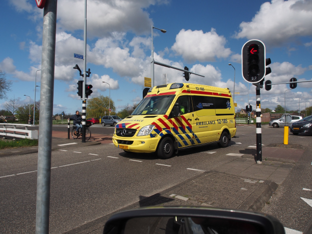 Ambulanz fährt auf einer Straße neben einer Ampel, mit einem Radfahrer im Vordergrund und Gebäuden, Bäumen und Laternen im Hintergrund.