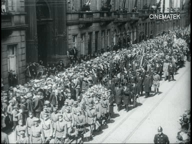 Schwarzes und weißes Foto einer Parade mit einer großen Menge Menschen, die eine Straße entlang gehen, einige halten Gewehre in den Händen, vor einem Gebäude.
