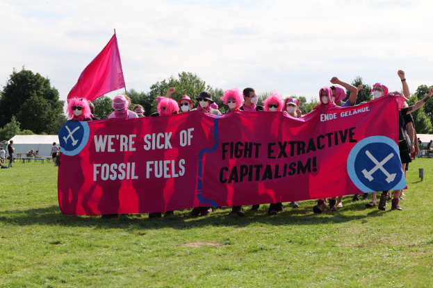 Gruppe von Menschen mit einer pinken Fahne, auf der "Wir haben genug von fossilen Brennstoffen, Kampf gegen den extraktiven Kapitalismus" steht, auf einer grünen Wiese mit Zelten, Bäumen und einem klaren blauen Himmel im Hintergrund.