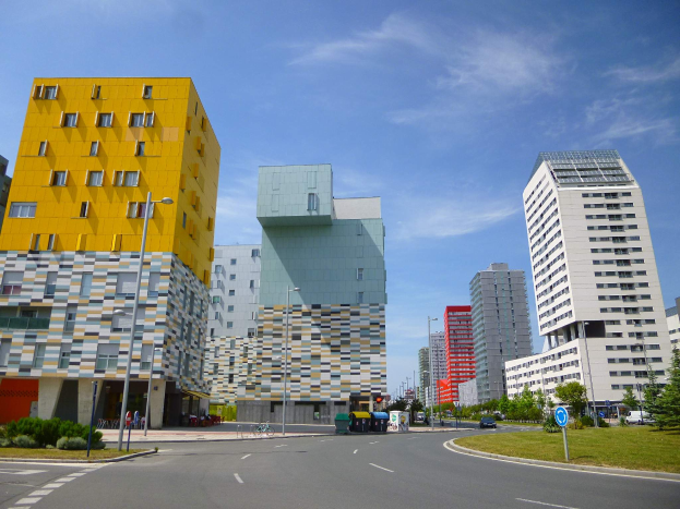 Eine Stadtstraße mit hohen, einzigartig gestalteten Universitätsgebäuden im Hintergrund, Straßenlaternen, Straßenlaternen, Kraftfahrzeuge, Informationsschilder, Büsche, Bäume und ein bewölkter Himmel.