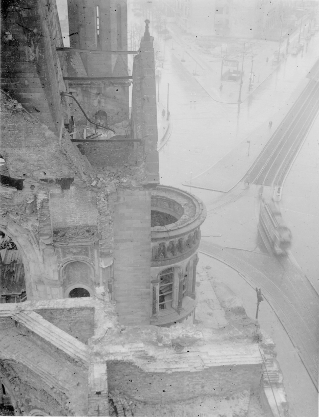 Schwarz-weiß-Foto einer Stadtstraße mit einem teilweise abgebrochenen Gebäude im Vordergrund, einigen Fahrzeugen auf der Straße und Strommasten auf der rechten Seite.
