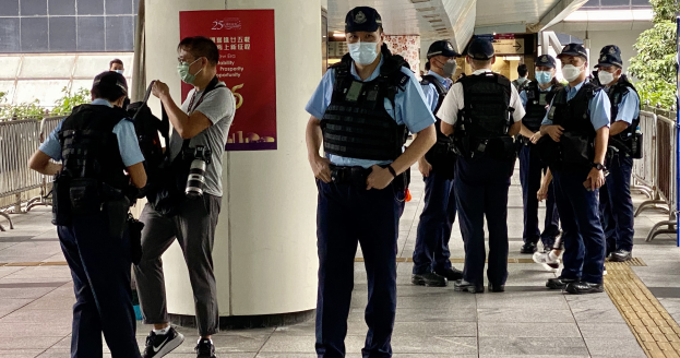 Polizisten in Uniform, mit Mützen und Mund-Nasen-Schutz stehen vor einem Gebäude, einige halten Kameras, mit einem Banner an einer Säule und Grün im Hintergrund.