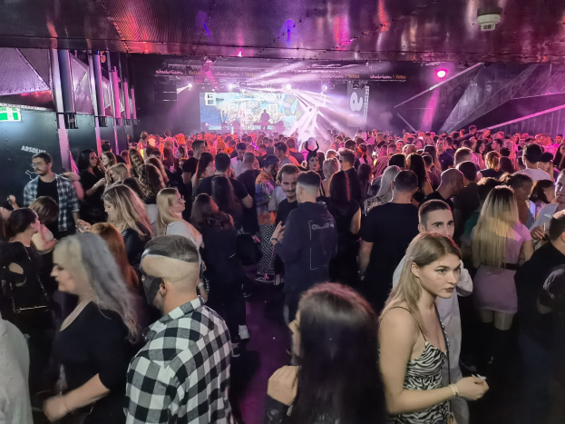 Eine große Menschenmenge in einer Diskothek mit Deckenbeleuchtung und Säulen, mit einem Bildschirm im Hintergrund, der als Absolut Nightlife-Event gekennzeichnet ist.