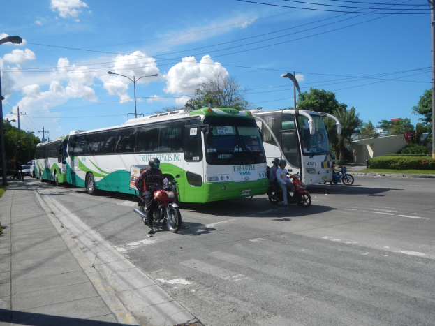 Ein grüner und weißer Shuttlebus steht am Straßenrand mit ein paar Motorrädern davor, ein Fußweg mit Gras und Pflanzen links daneben und Gebäude, Bäume, Laternenpfähle und einen klaren blauen Himmel im Hintergrund.