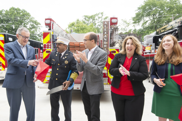 A group of people clapping and smiling at a ribbon cutting ceremony for the Alexandria Fire Department, with two individuals holding scissors and a red ribbon in front of a fire truck, set against trees, wires, and a clear blue sky.
