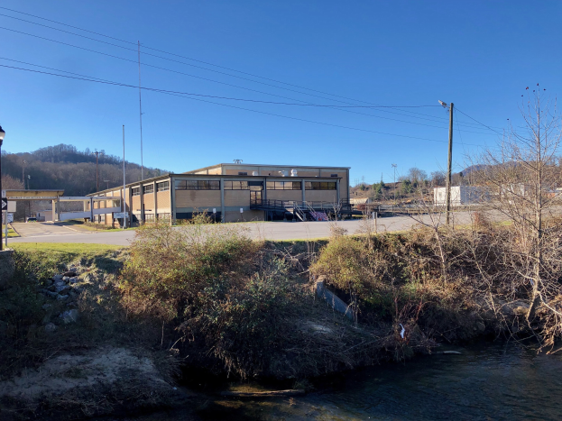 Ein Fluss fließt durch ein grünes Feld neben einem Gebäude, mit Bäumen, Pflanzen, Gras, Steinen, Straßenlaternen, Strommasten, Kabeln, Hügeln und einem klaren blauen Himmel im Hintergrund; das Gebäude befindet sich an der Stelle einer geplanten Wasseraufbereitungsanlage.